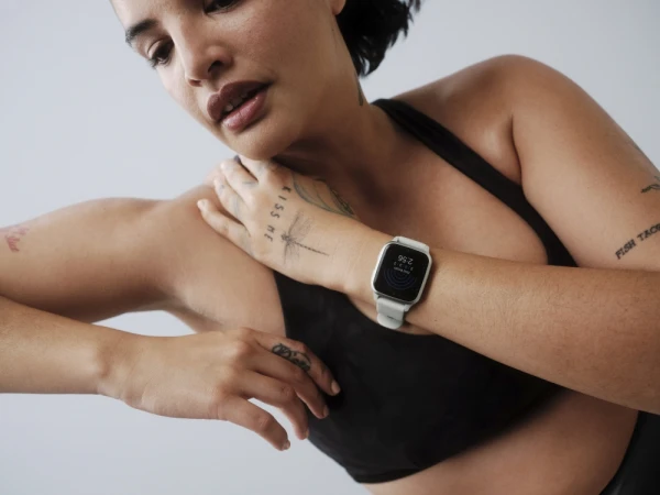 Woman wearing a black sports bra and a smart watch with white band, resting her hand on her shoulder in a bright, neutral setting.