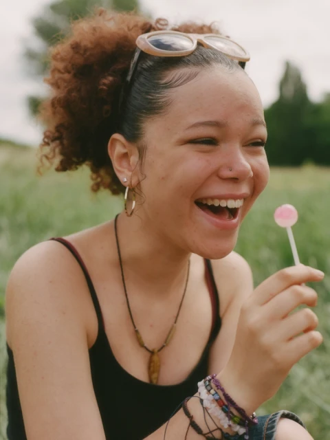 Portrait of a smiling young woman with curly hair outdoors holding a lollipop in a grassy field, wearing sunglasses and jewelry.