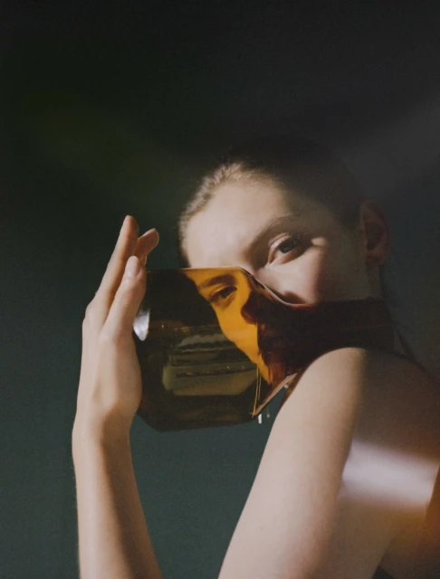 A woman holds a transparent, amber cube reflecting her face in front of her face in a studio with diffused lighting.