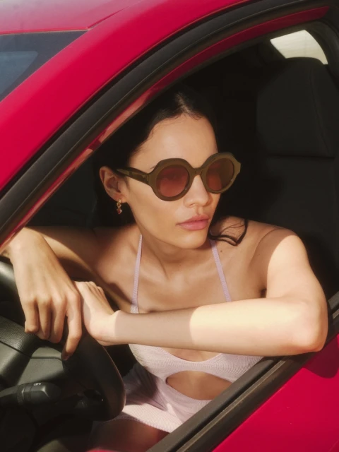 Brunette woman with sunglasses sitting in a red car, wearing a light purple dress and gold earrings with steering wheel visible.