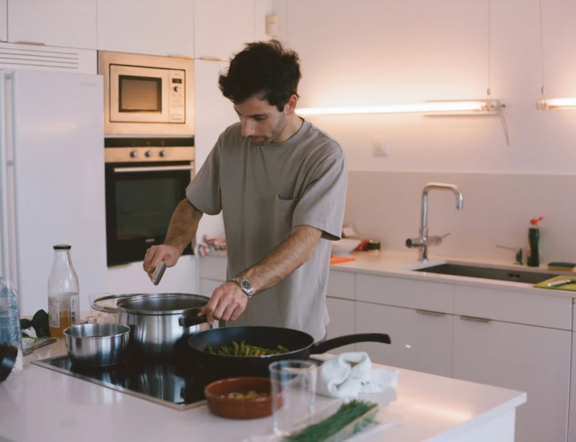 Man cooking in a bright, modern kitchen with stainless steel appliances, preparing food in a pan and pot on a white countertop.
