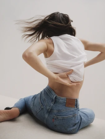 Woman wearing a white tank top and blue jeans sitting on a white background with hair blowing