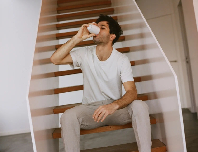 Adult male in white t-shirt sits on wooden stairs indoors drinking from a white bottle, neutral color palette. Minimalist interior design.