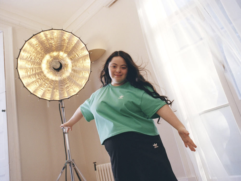 Smiling woman in green Adidas t-shirt and black skirt posing in front of a large studio light and white curtain, indoor setting.