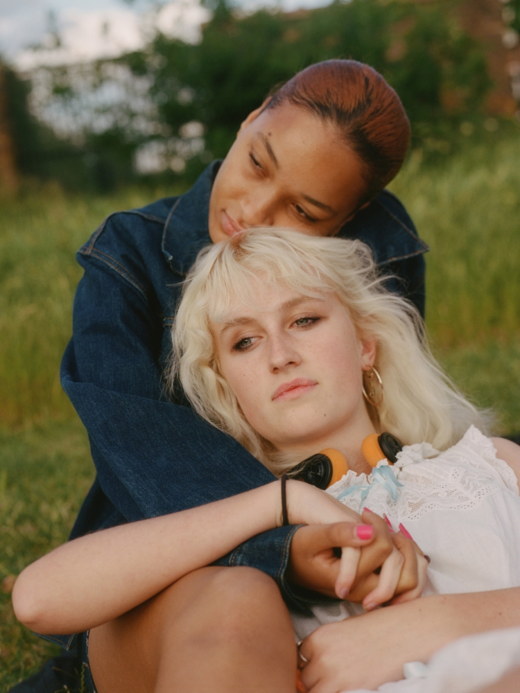 Two women embrace in an outdoor setting with grass and greenery. One woman with short brown hair rests her head on the other woman.