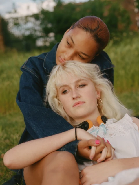 Two women embrace in an outdoor setting with grass and greenery. One woman with short brown hair rests her head on the other woman.