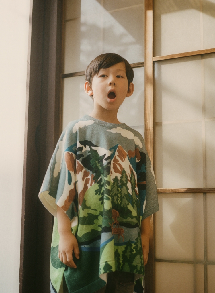 Young child in a printed poncho stands in front of a window, looking up with an open mouth and a surprised expression indoors.