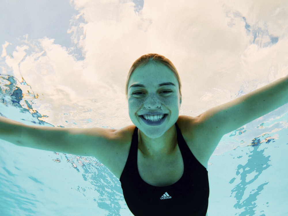 Smiling blonde woman in a black swimsuit swims underwater in a pool, looking up towards the sunlight and cloudy sky above the surface.