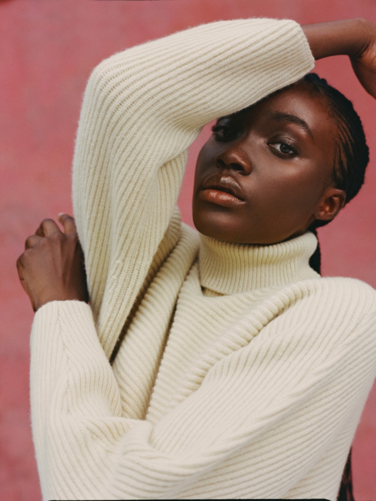 Portrait of a young Black woman in a cream turtleneck sweater against a pink backdrop, looking at the camera with arms raised.
