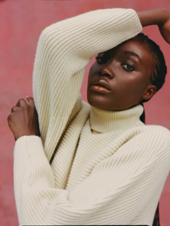 Portrait of a young Black woman in a cream turtleneck sweater against a pink backdrop, looking at the camera with arms raised.