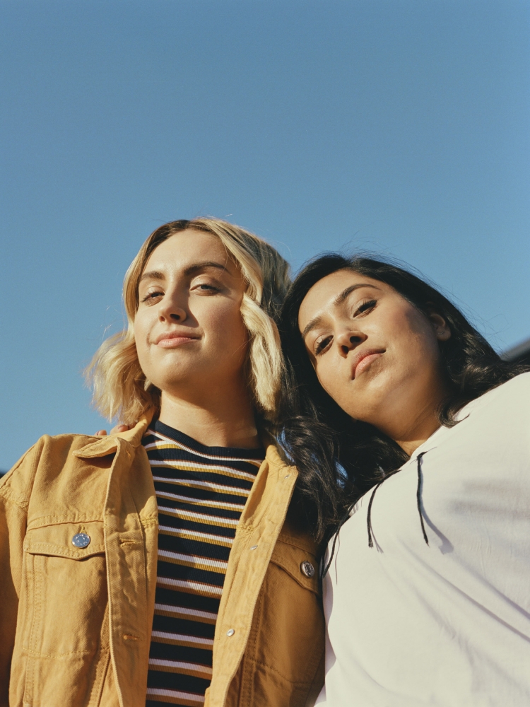 Two young women standing side by side outdoors against a clear blue sky, photographed from a low angle by Ana Cuba.