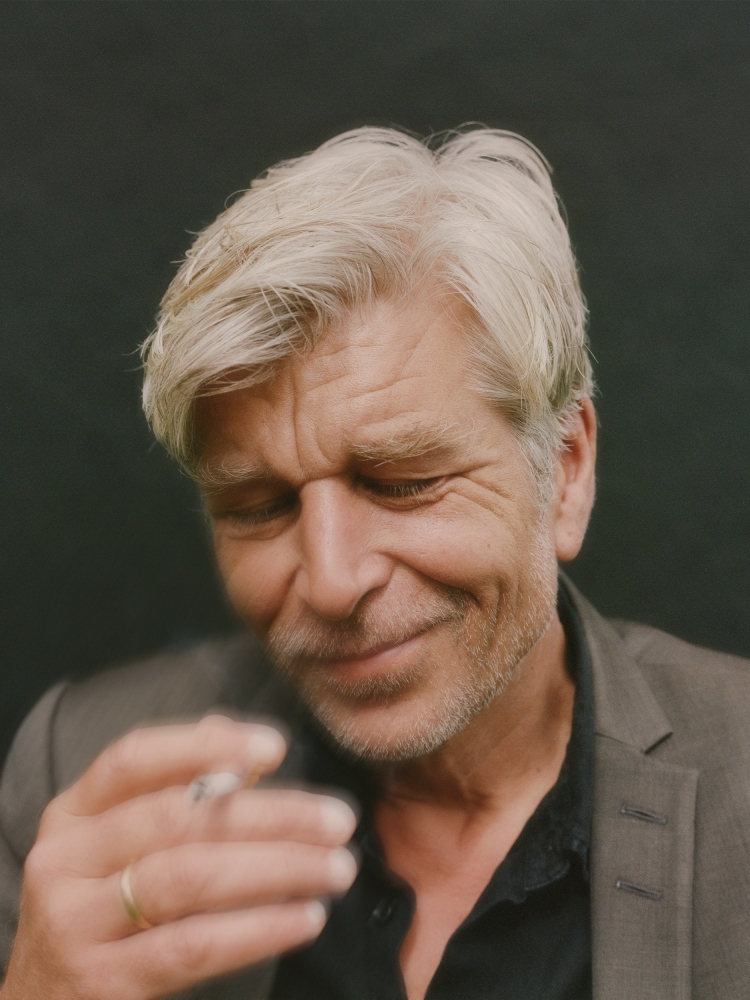 Portrait of a mature adult male with graying hair wearing a suit jacket and smoking a cigarette indoors against a dark backdrop.