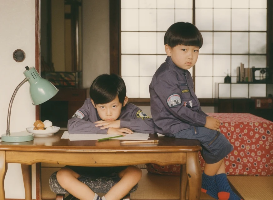 Two young boys pose indoors near a wooden desk and Japanese-style sliding doors, with one child sitting on the desk and another sitting below.