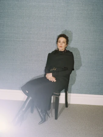 Portrait of a seated woman wearing a black dress and boots in front of a blue textured wall with an off-camera light flare.