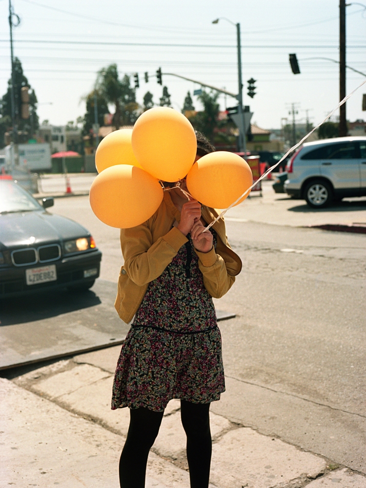 A person stands on a sidewalk holding four yellow balloons in front of their face with cars and traffic lights in the background.