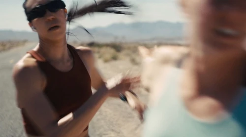Woman with sunglasses running on road in desert landscape with another person blurred in foreground and mountains in background.