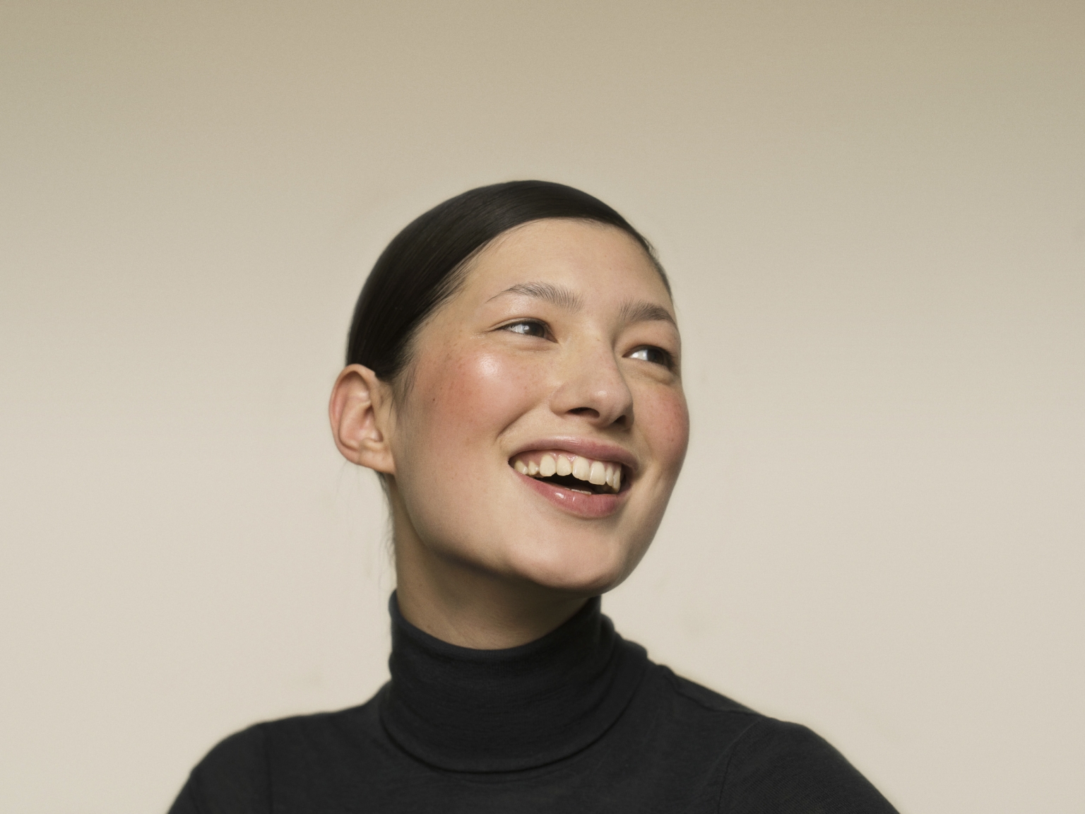 Close-up studio shot of a young Asian woman with a happy expression, wearing a black turtleneck sweater against a neutral background.