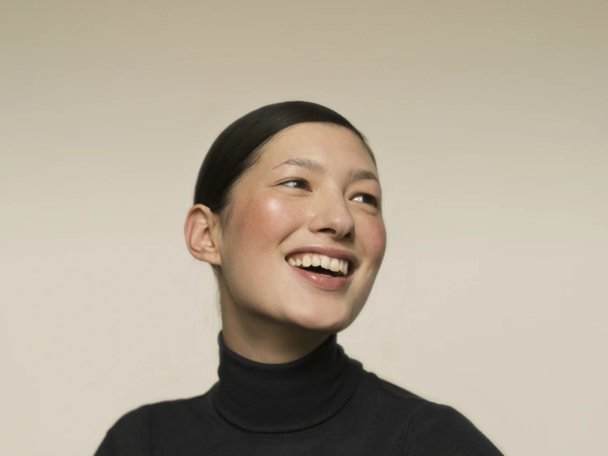 Close-up studio shot of a young Asian woman with a happy expression, wearing a black turtleneck sweater against a neutral background.