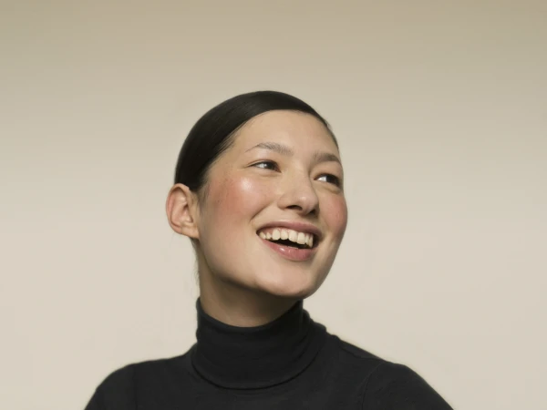 Close-up studio shot of a young Asian woman with a happy expression, wearing a black turtleneck sweater against a neutral background.