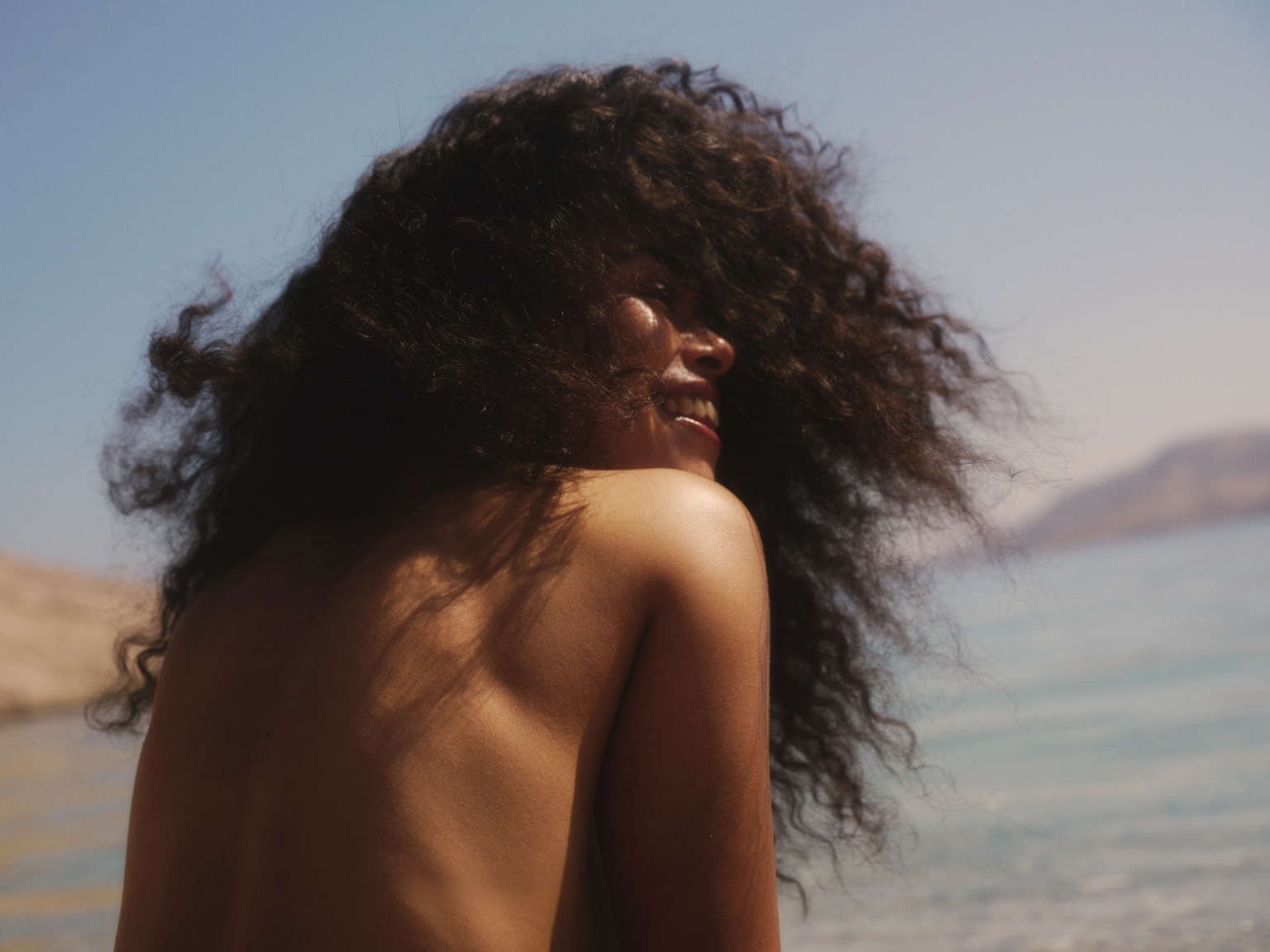 Woman with long dark curly hair looking back over her shoulder outdoors near water under a clear sky.