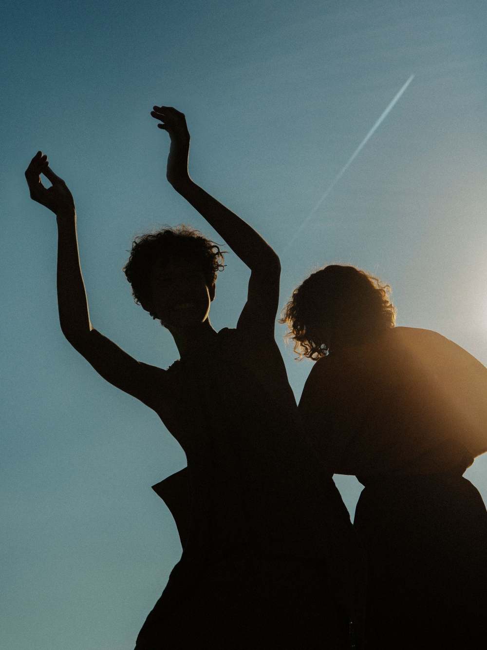 Silhouette of two people against a clear blue sky at daytime, one with arms raised, captured in an artistic and minimalist style.