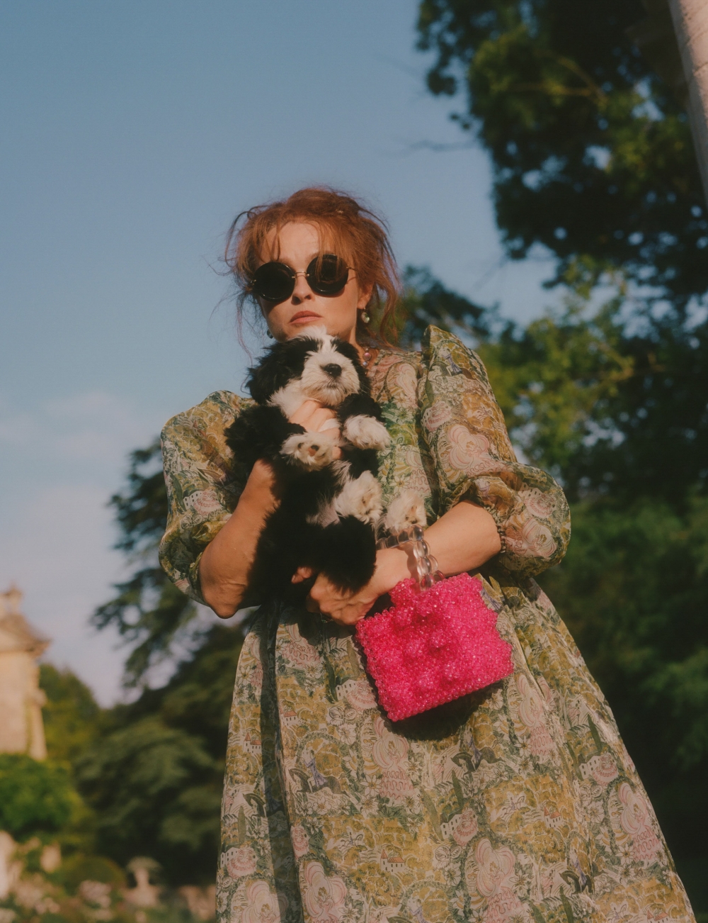 Adult woman holding a puppy outdoors in a paisley dress with a pink bag and sunglasses near green trees under a blue sky.