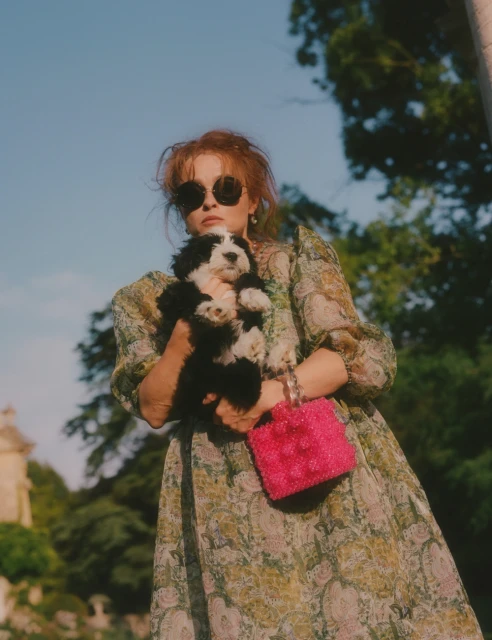 Adult woman holding a puppy outdoors in a paisley dress with a pink bag and sunglasses near green trees under a blue sky.
