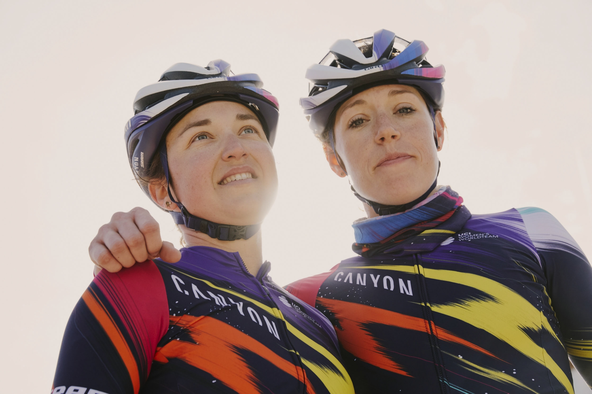 Two female cyclists in matching racing kits and helmets pose together in a low-angle outdoor shot with bright, diffused lighting.