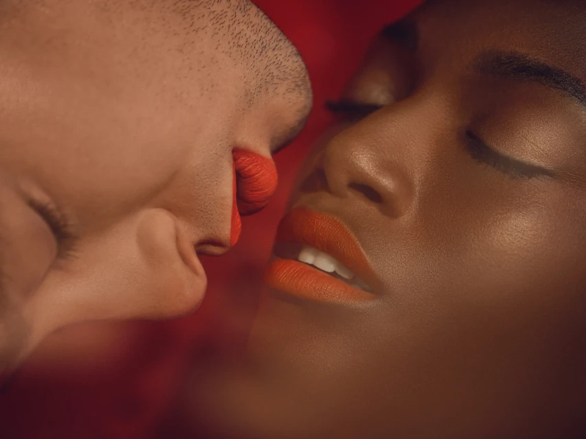 Close-up of faces of a fair-skinned man and a dark-skinned woman in muted lighting with a red backdrop, both wearing red lipstick.