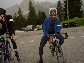 Cyclist in a blue jacket and white helmet laughing while leaning on a road bike outdoors in a mountainous parking area.