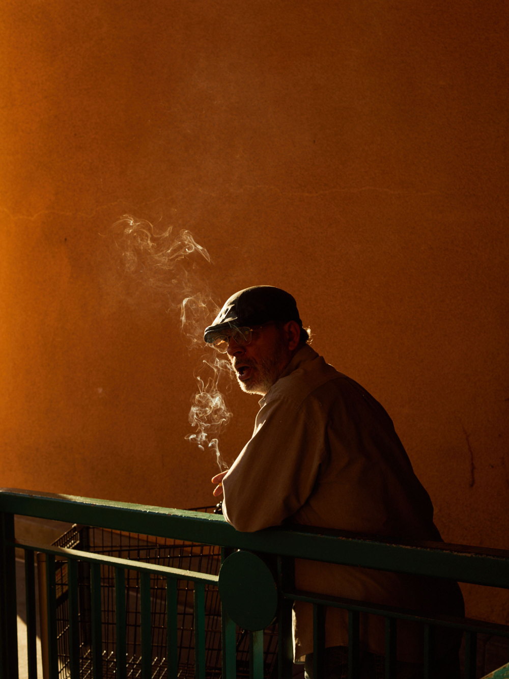 An adult man in a cap stands against an orange wall, smoking, with a railing in the foreground, creating an introspective and atmospheric scene.