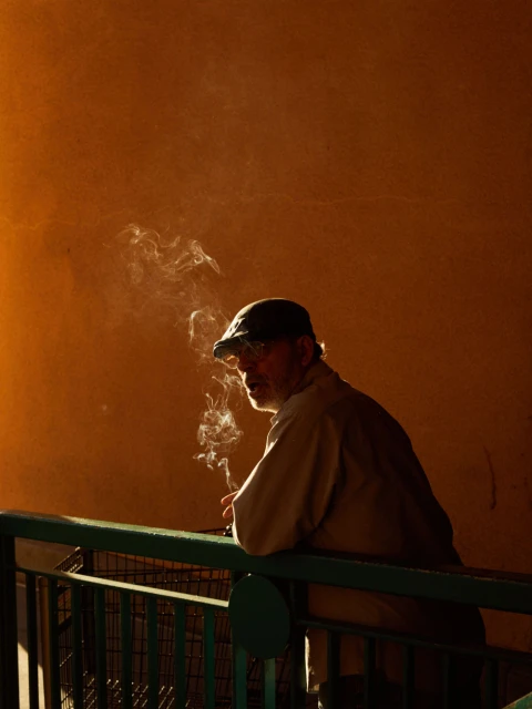 An adult man in a cap stands against an orange wall, smoking, with a railing in the foreground, creating an introspective and atmospheric scene.