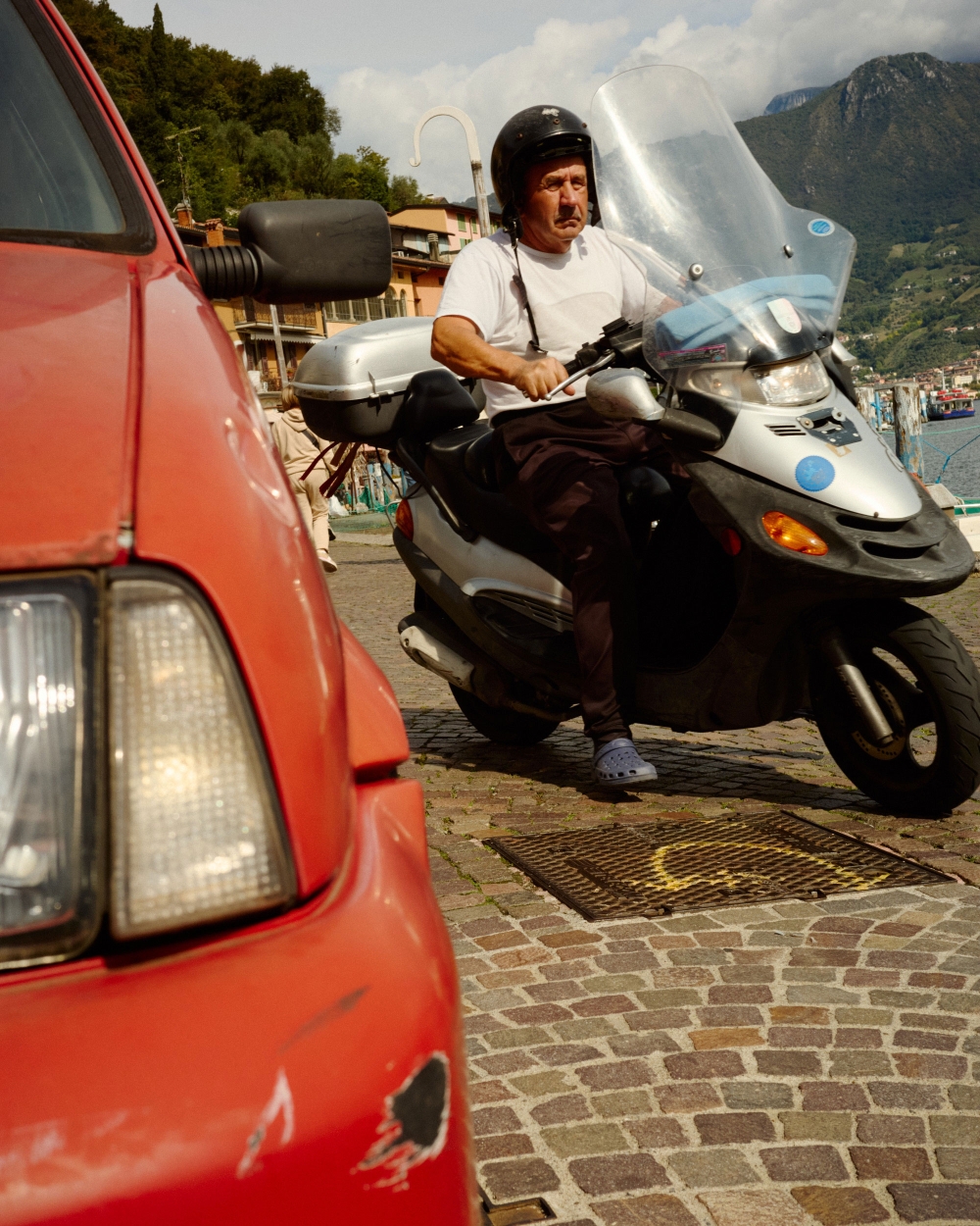 Adult male riding a silver scooter on a cobblestone road near a body of water, wearing a helmet. Red car visible to the left.