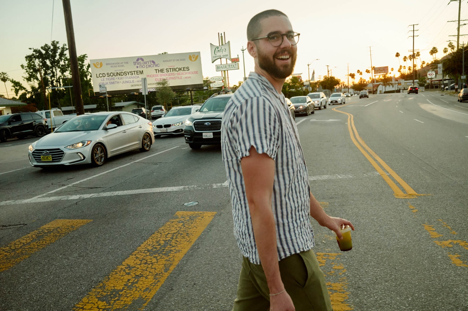Adult male with a smile crossing a city street with a drink. Cars are stopped in the background and the sun is setting.