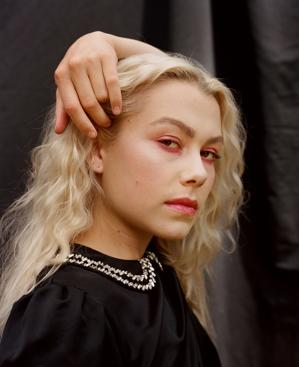 Close-up portrait of a young adult female with blonde wavy hair, wearing a black dress with a jeweled collar, posing against a dark background.
