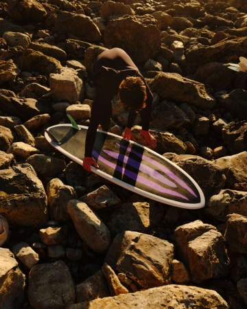 Person in wetsuit bends over surfboard on rocky beach in outdoor shot with natural light.