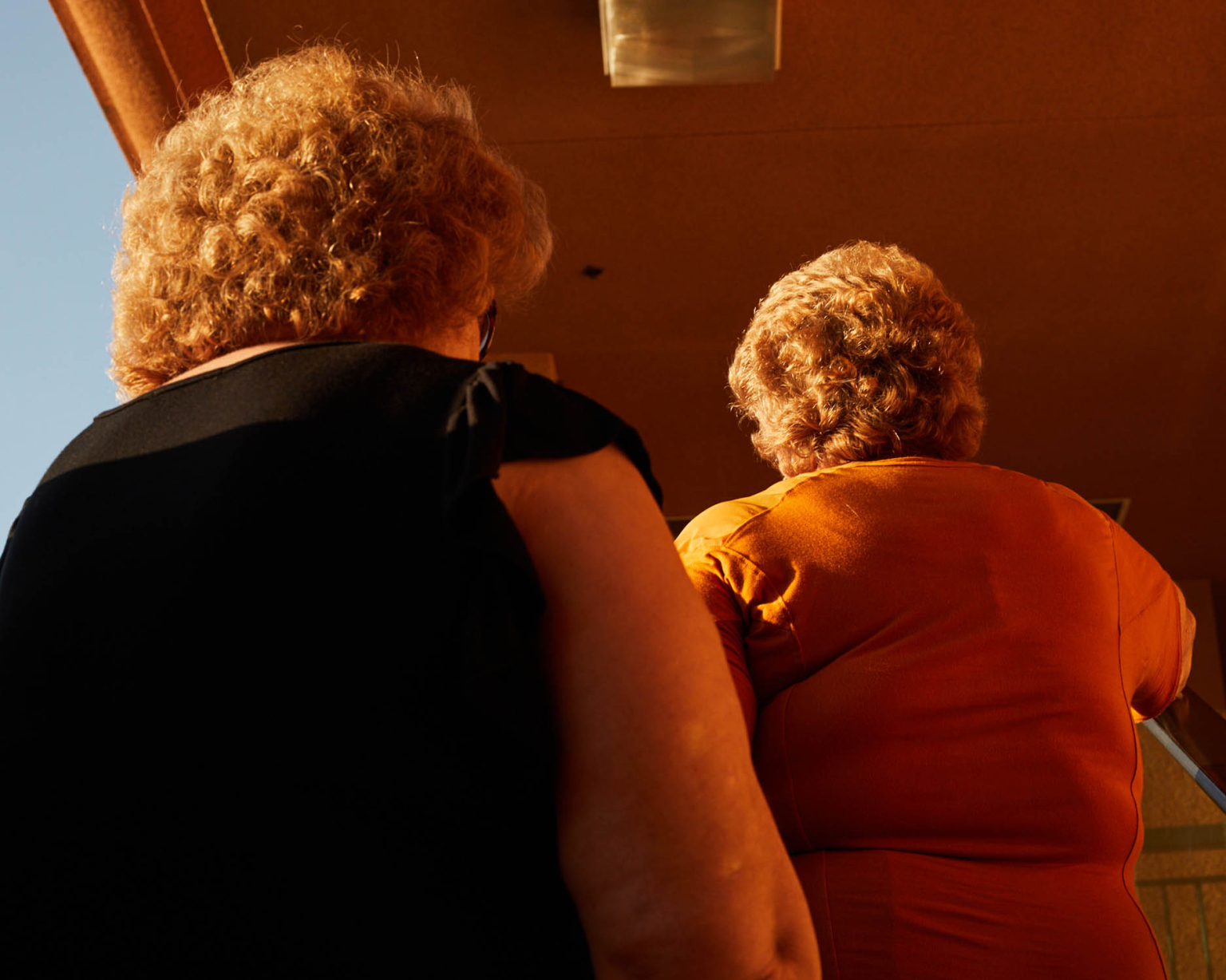 Back view of two elderly women with curly red hair, one in black, one in orange, standing indoors with warm lighting.