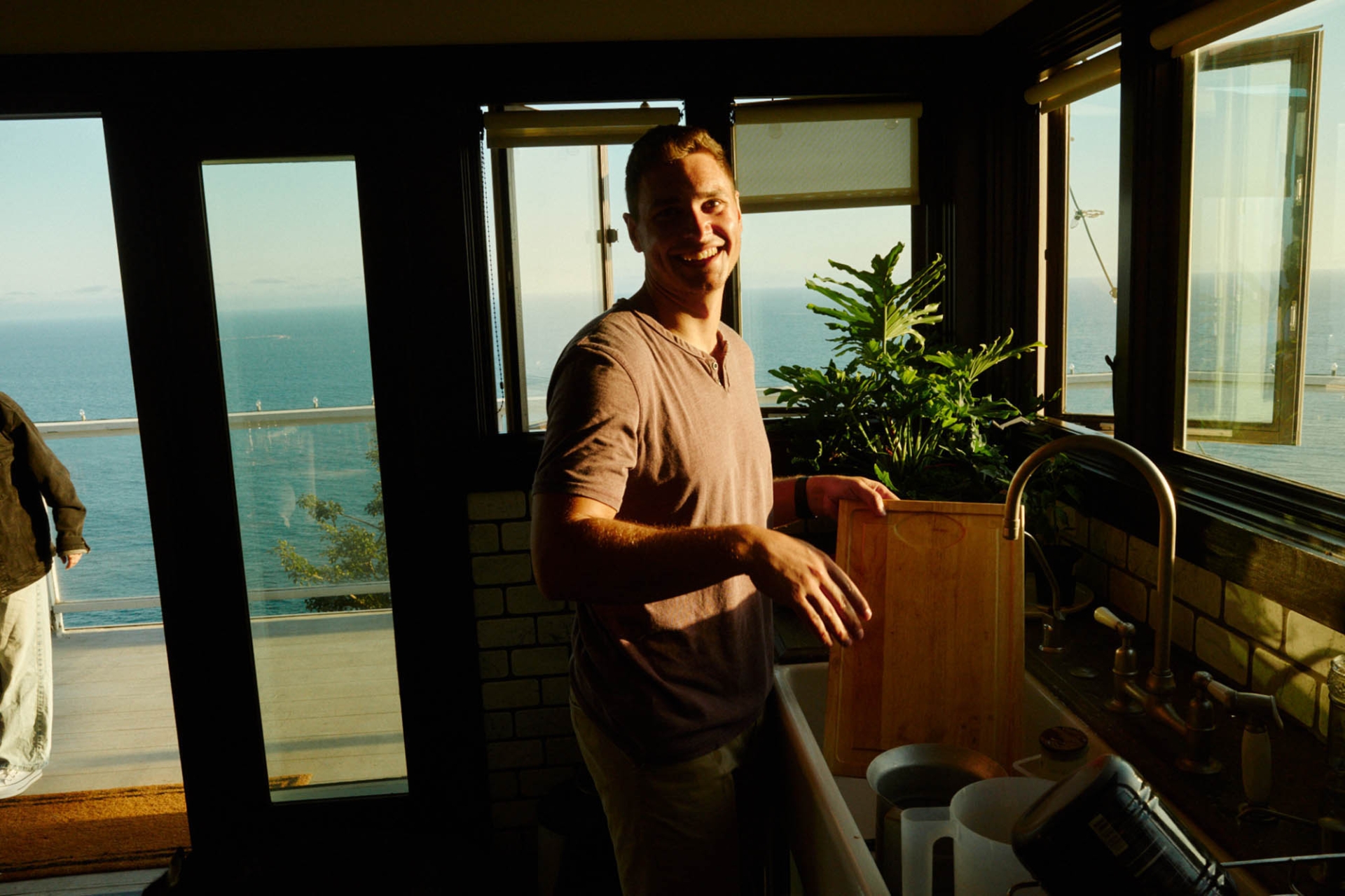 Man smiles in an indoor kitchen setting; ocean view visible through the windows. Sunlight brightens the room; indoor plants add a touch of nature.