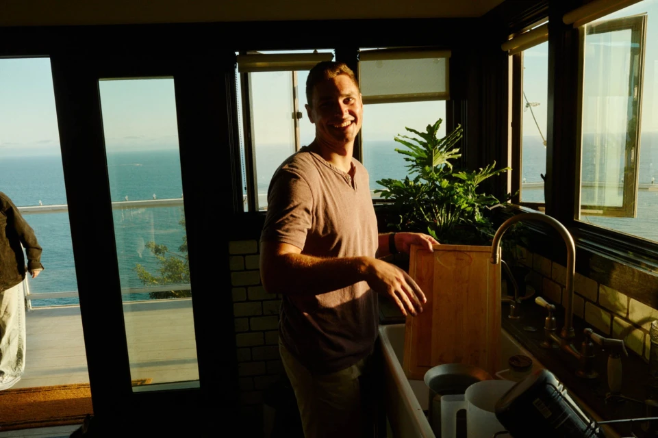 Man smiles in an indoor kitchen setting; ocean view visible through the windows. Sunlight brightens the room; indoor plants add a touch of nature.