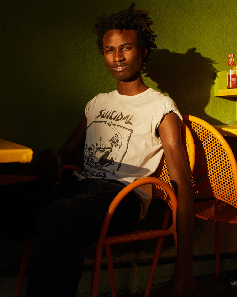 A Black man wearing a Suicidal Tendencies shirt poses indoors in a yellow and orange color-blocked interior setting, with green walls and sunlight.