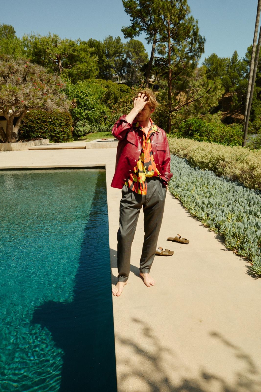 Man in casual attire stands barefoot near a pool on a sunny day, with lush greenery in the background, creating a relaxed outdoor scene.