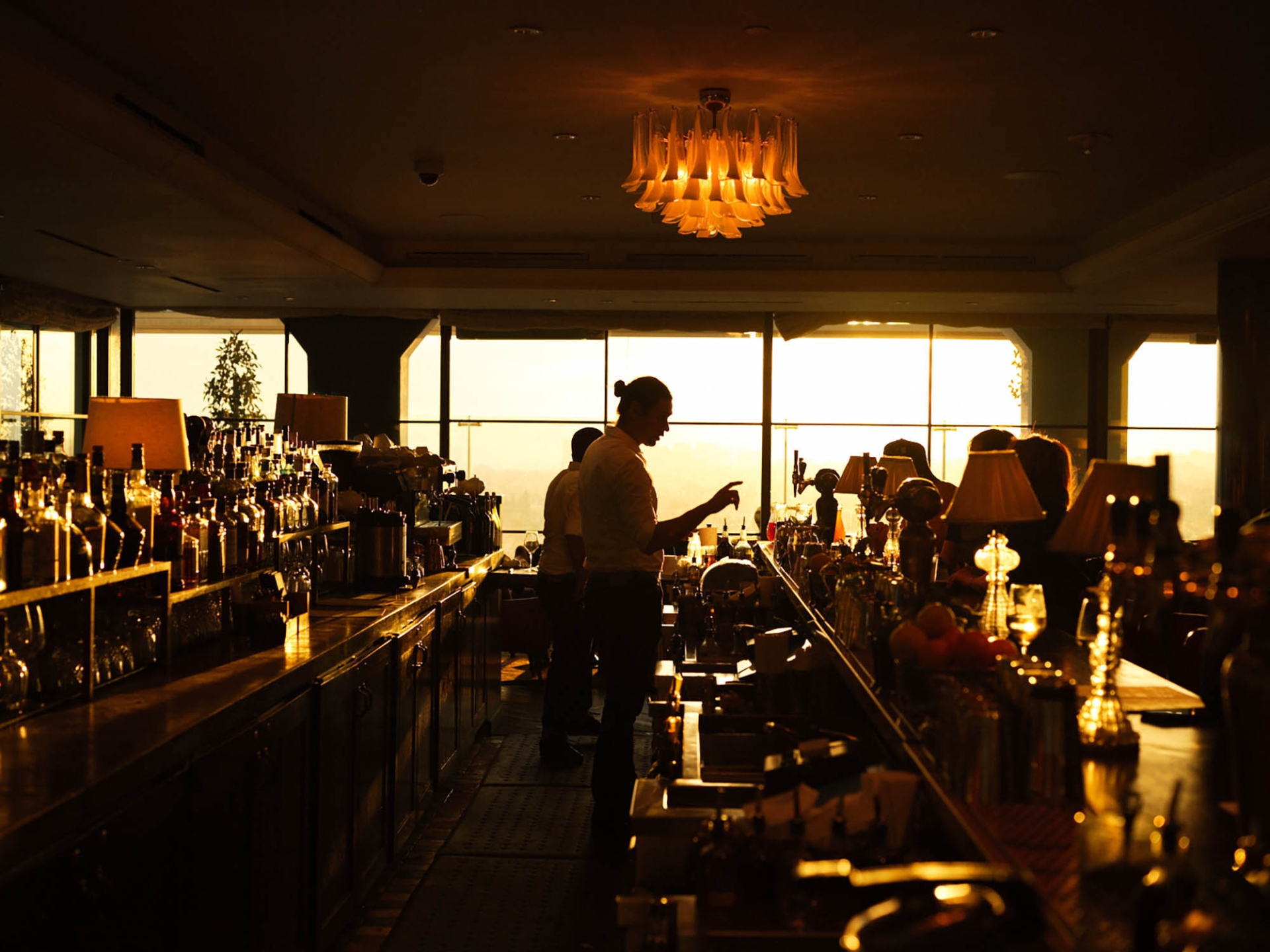 Silhouette of bartenders working at a bar with many bottles and glasses against sunset background, warm lighting, and decorative chandelier.