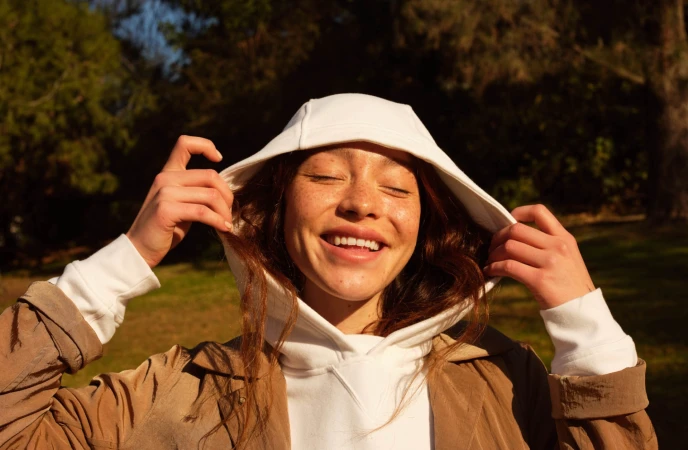 Close-up portrait of a smiling woman with closed eyes wearing a white hoodie and a brown jacket outdoors in Los Angeles.