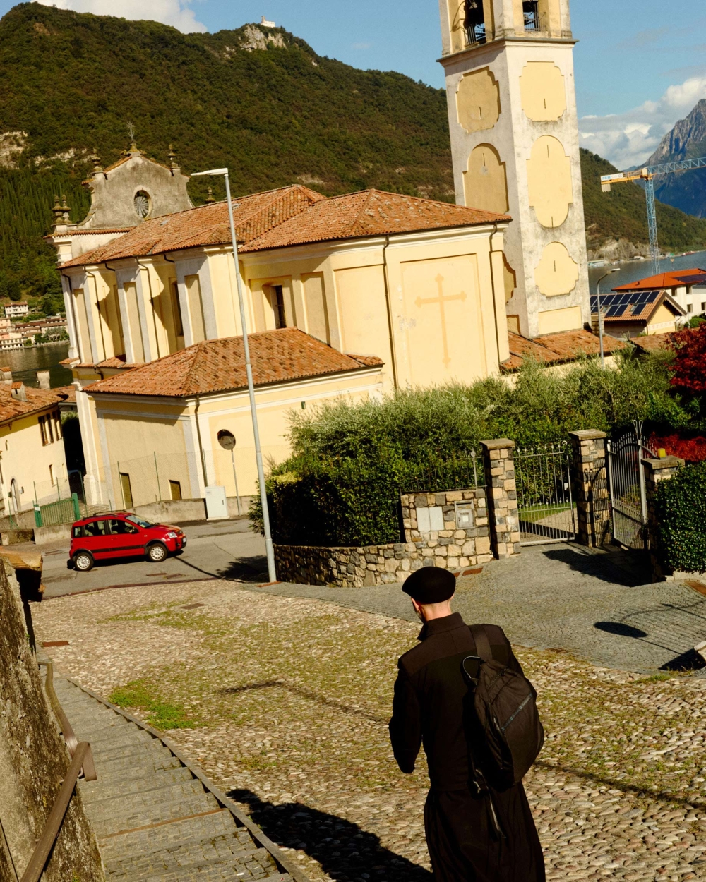 View of a cleric walking on a cobblestone path near a church and a lake surrounded by mountains on a sunny day, with a red car in the background.