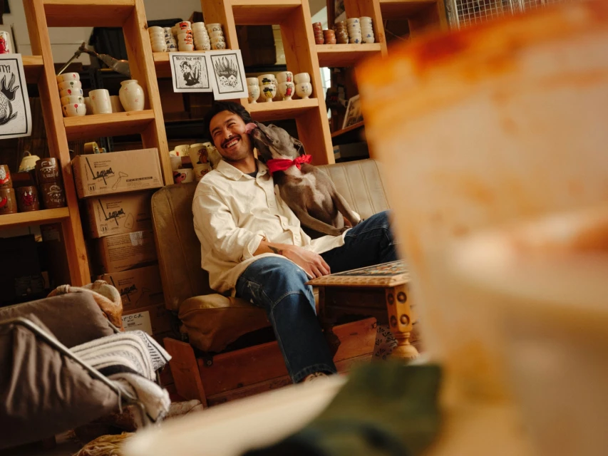 Adult man smiles as a dog licks his face while seated on a couch in a room filled with shelves and ceramic mugs.