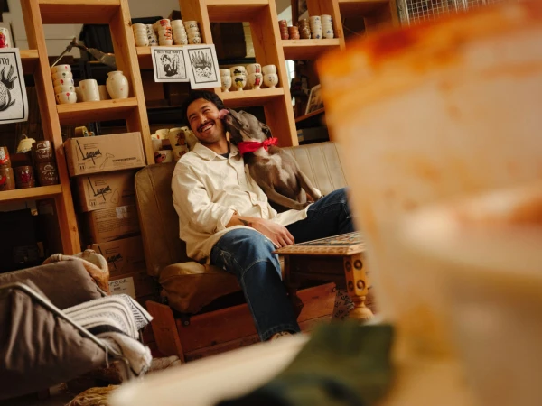 Adult man smiles as a dog licks his face while seated on a couch in a room filled with shelves and ceramic mugs.