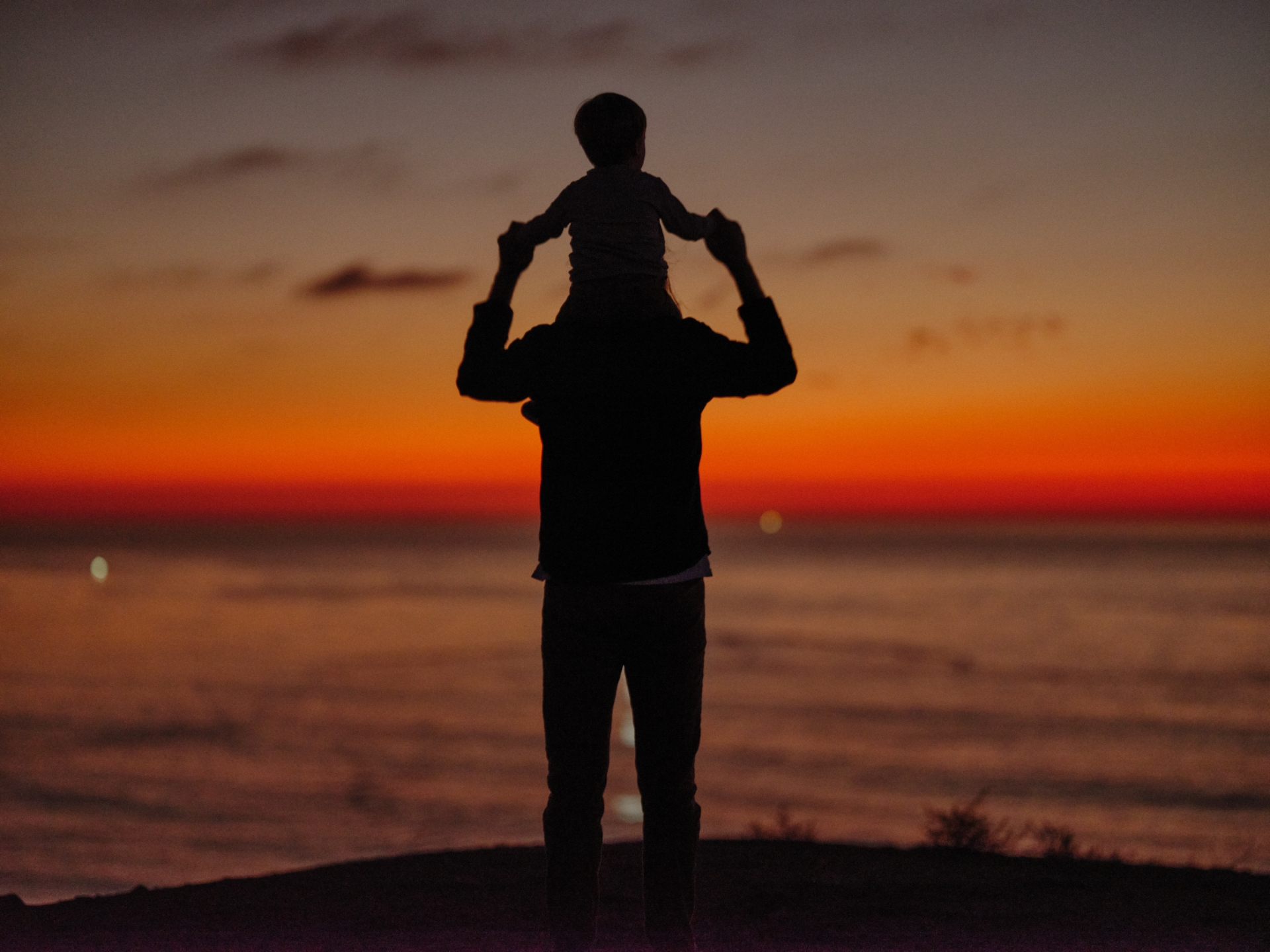 Silhouette of a parent holding a child on their shoulders at sunset, overlooking the ocean with a vivid orange and red sky.