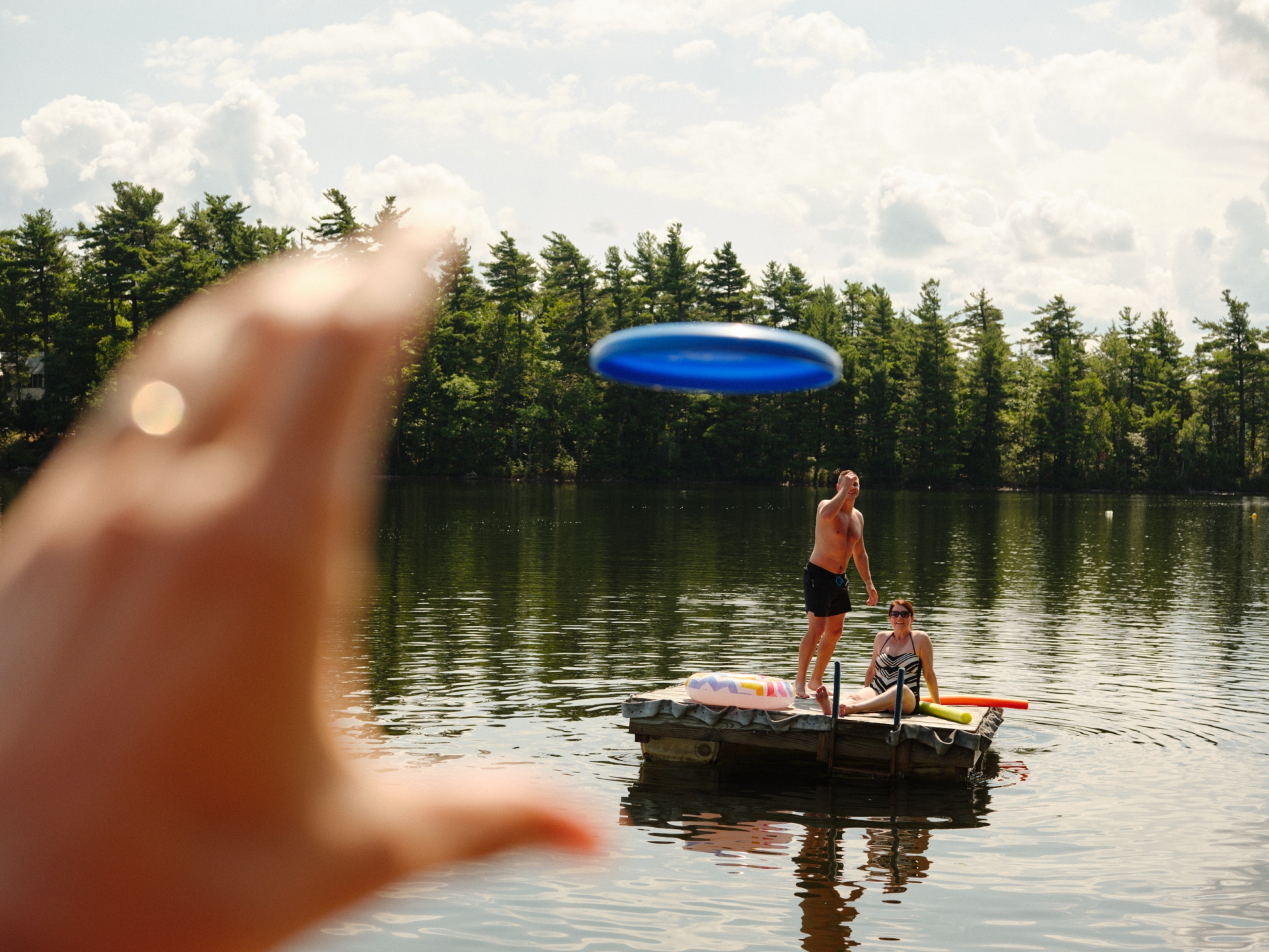 A man throws a blue disc while standing on a raft with a woman in the water, with a blurred hand in the foreground.