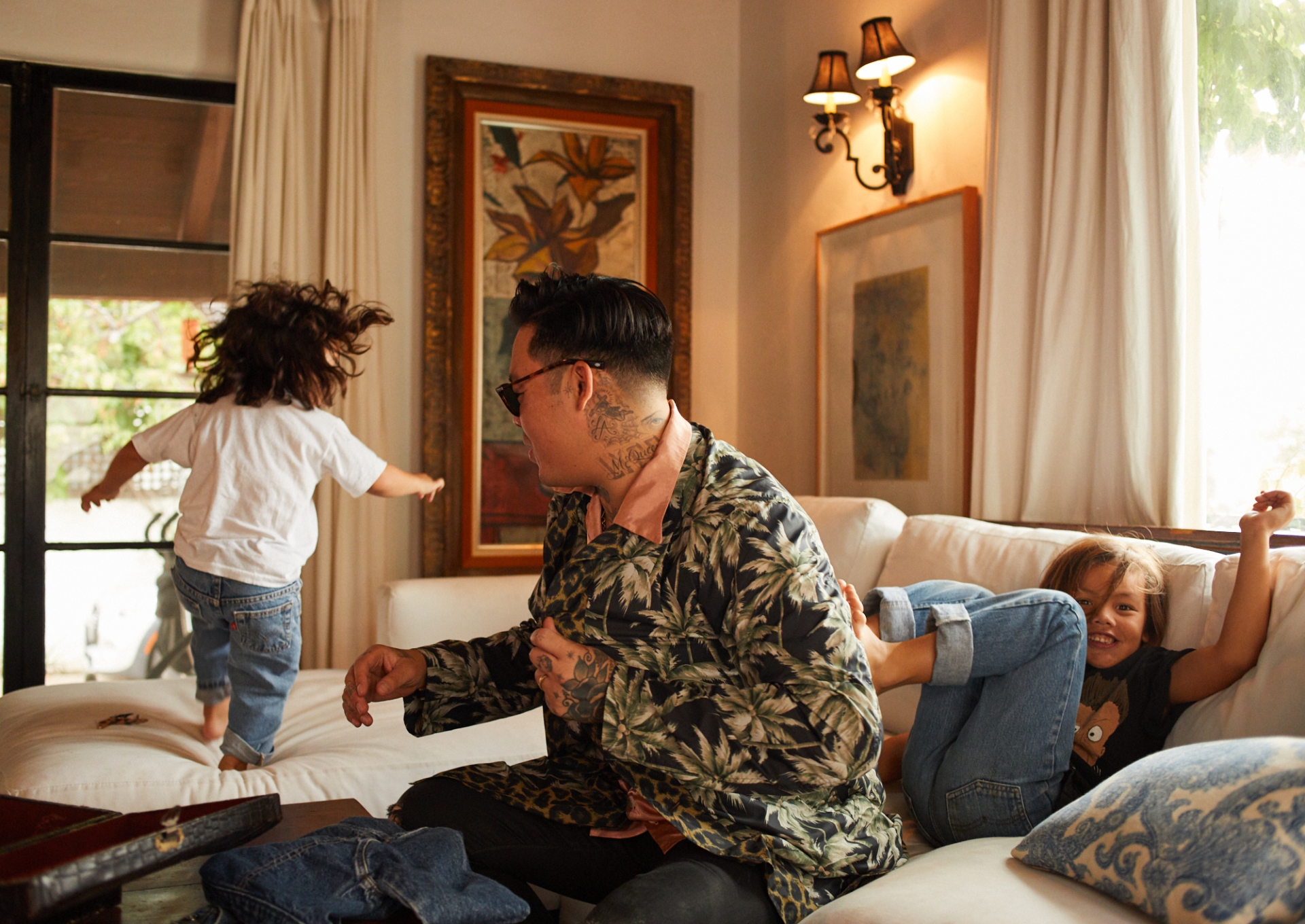 An adult and two children interact on a white sofa in a warmly lit indoor living space, featuring art, decorative lighting, and natural light.