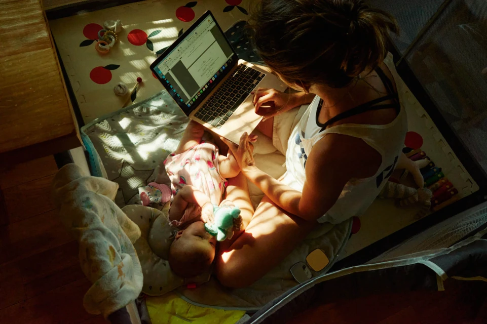 Woman works on a laptop while sitting on the floor with her baby in the sunlight, in an indoor setting near a playpen.